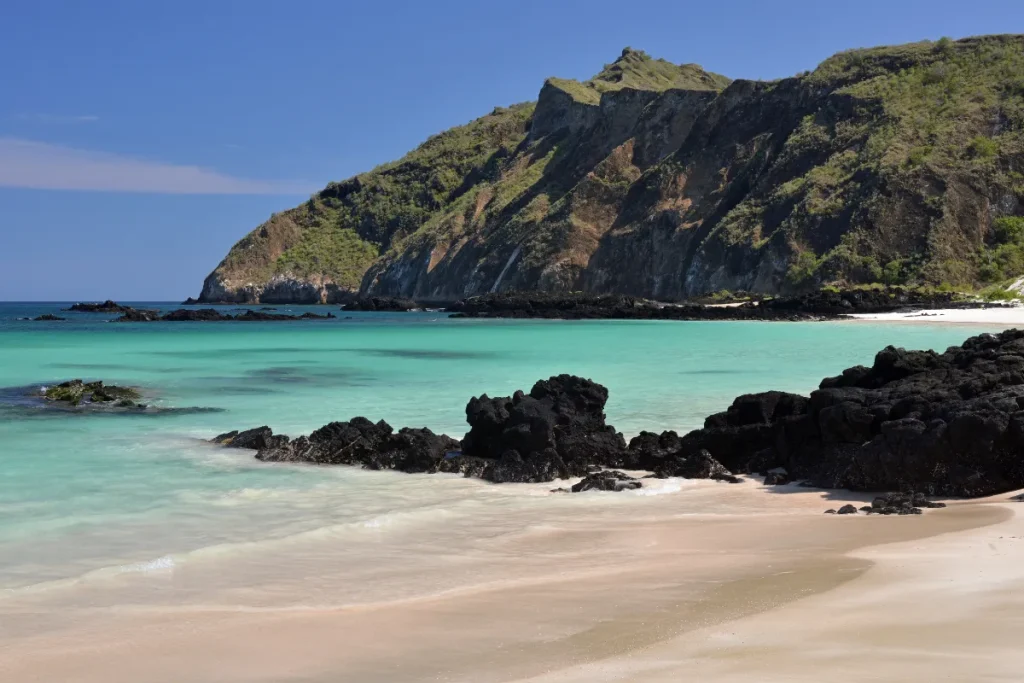 Praia de areia clara com rochas escuras e mar turquesa nas Ilhas Galápagos, com colinas verdes ao fundo.