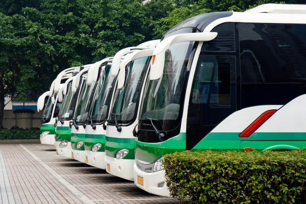 Fila de veículos elétricos modernos do transporte público estacionados lado a lado em uma área pavimentada com árvores ao fundo.