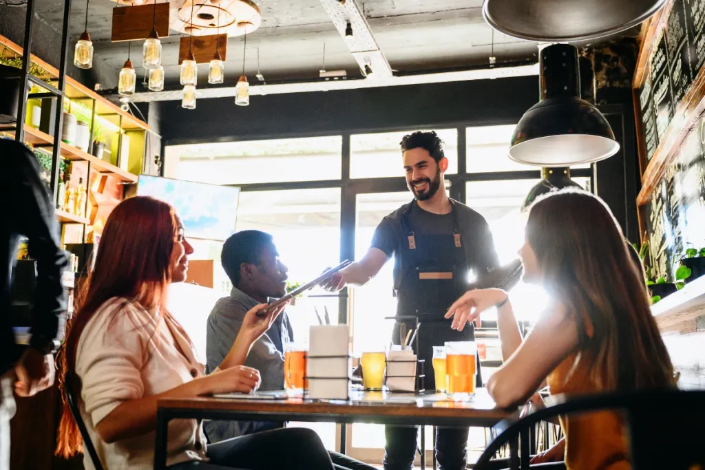 Grupo de pessoas sentado em uma mesa de restaurante enquanto um garçom serve bebidas em um ambiente interno iluminado.