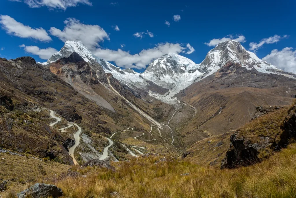 Paisagem da Cordilheira dos Andes com montanhas altas, trilha sinuosa e picos nevados ao fundo.
