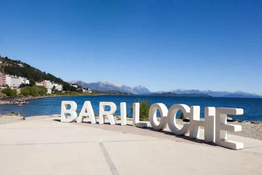 Letreiro com a palavra Bariloche em uma área aberta próxima ao lago Nahuel Huapi com montanhas ao fundo.