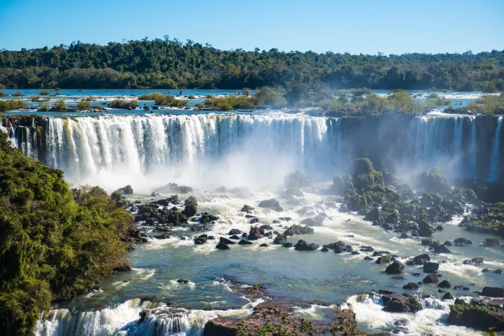 Vista panorâmica das Cataratas do Iguaçu com várias quedas d’água cercadas por vegetação na fronteira entre Brasil e Argentina.
