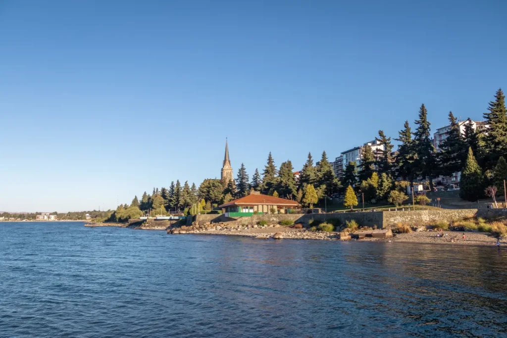 Orla do lago Nahuel Huapi com árvores, construções e igreja ao fundo no centro de Bariloche.