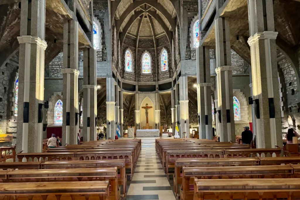 Interior da Catedral de Bariloche com bancos de madeira, altar ao fundo e vitrais coloridos nas paredes.