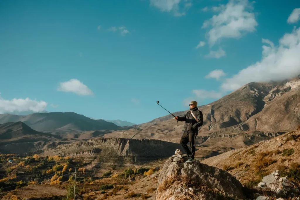 Homem em pé sobre uma rocha usando bastão de selfie em paisagem montanhosa ampla.