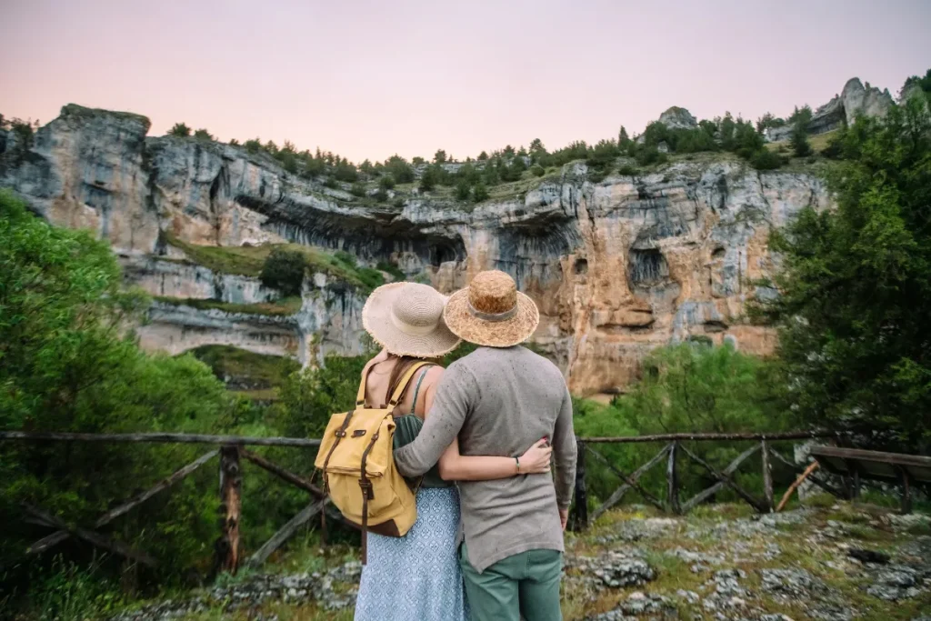 Casal usando chapéus e mochila abraçado enquanto observa formações rochosas em região montanhosa.