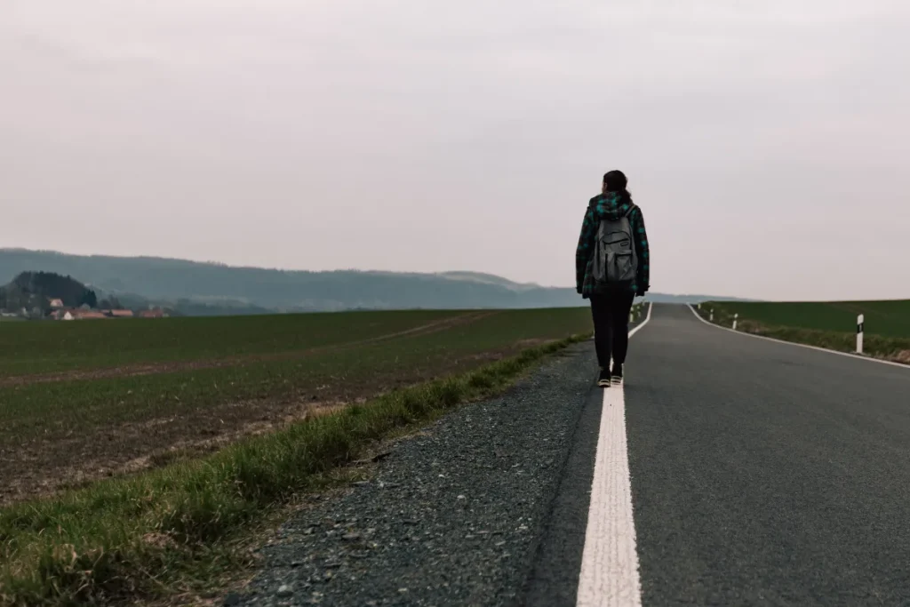 Pessoa com mochila caminhando sozinha pelo acostamento de uma estrada em área rural.