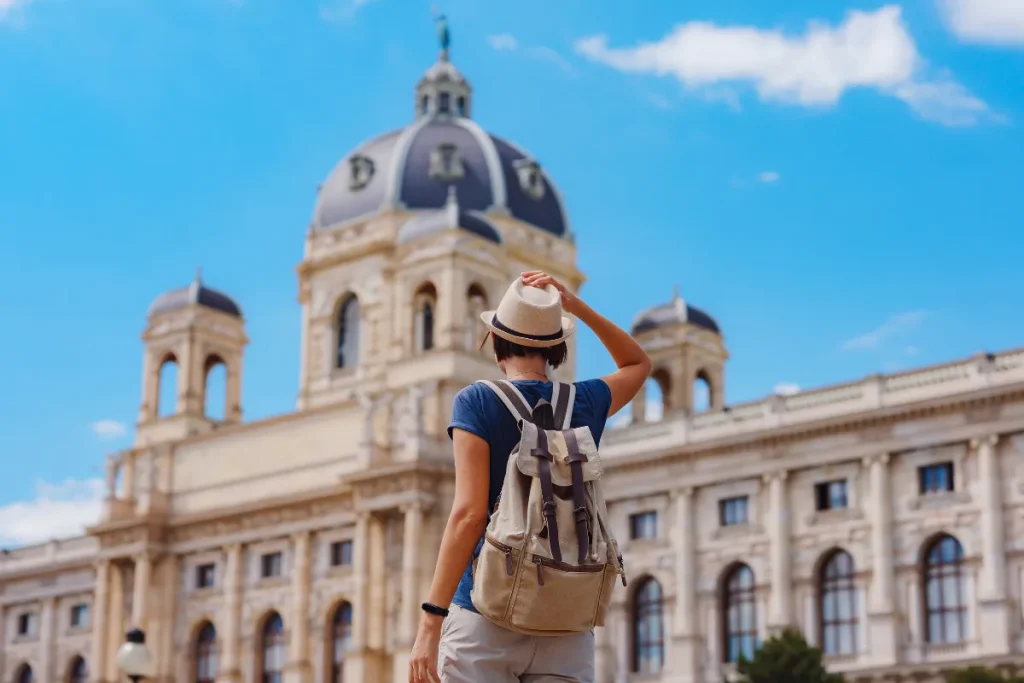 Mulher com mochila e chapéu observando prédio histórico com cúpula em praça urbana.