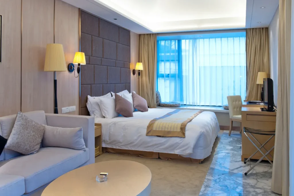Quarto de hotel com cama grande, sofá, mesa de trabalho e janela ampla com cortinas