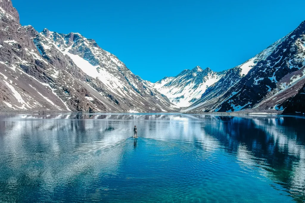 Lago cercado por montanhas nevadas com reflexo da paisagem na água e uma pessoa ao centro