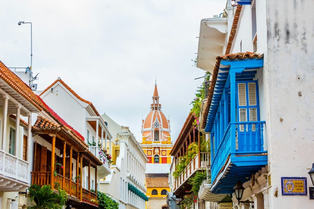 Rua histórica de Cartagena das Índias com sacadas coloniais coloridas e torre da catedral ao fundo.