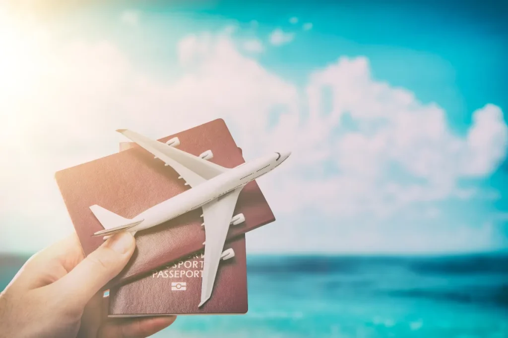 Quanto custa viajar para Bariloche: valores de hotel, comida e passeios em reais Mão segurando passaporte com avião em miniatura em frente ao mar e céu azul