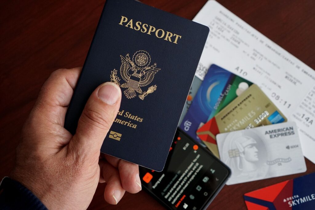 Pessoa segurando passaporte ao lado de cartões bancários, celular e cartão de embarque, representando viagens internacionais e documentação de viagem.