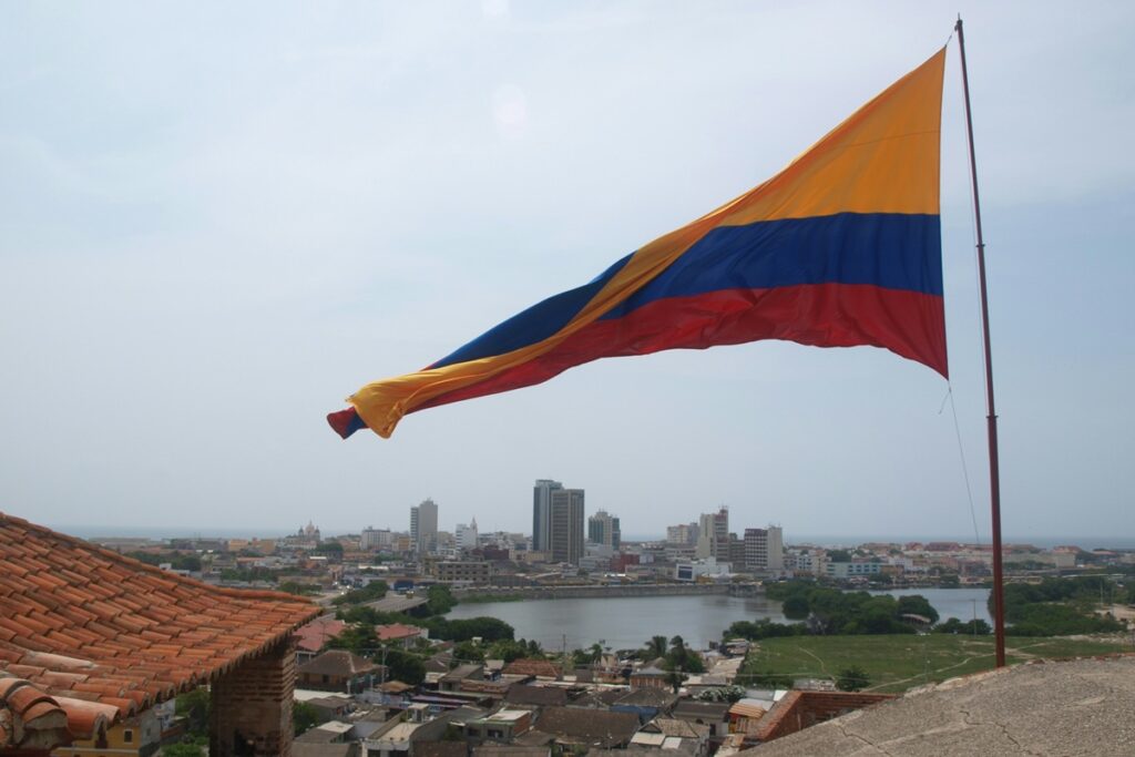 Bandeira da Colômbia hasteada sobre paisagem urbana com telhados coloniais e prédios ao fundo.