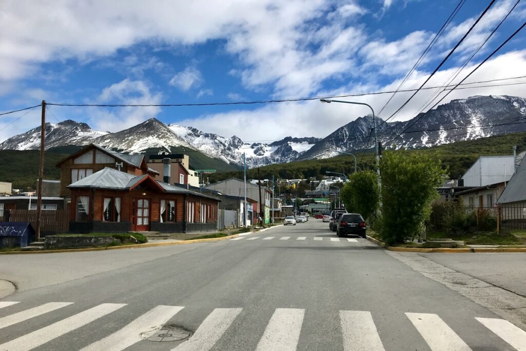 Rua de Ushuaia com casas, faixa de pedestres e montanhas nevadas ao fundo na Patagônia Argentina.