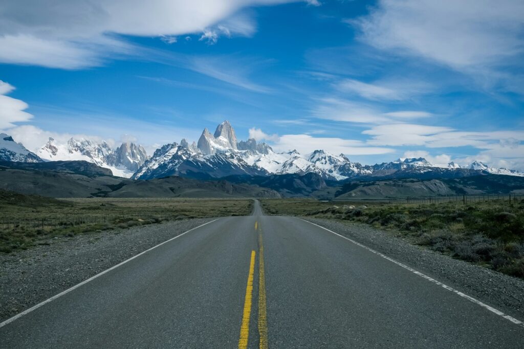 Estrada vazia em direção ao Monte Fitz Roy, na Patagônia, com montanhas nevadas e céu azul.