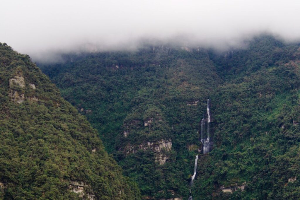 Cachoeira estreita descendo por uma montanha coberta de vegetação, parcialmente encoberta por nuvens baixas.